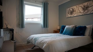 Ein Schlafzimmer mit einem Bett, einem Nachttisch, einer Lampe, einem Fenster mit Vorhängen und einem Wandbild.