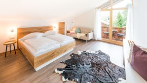 A bedroom with a wooden bed, a small side table, a sofa, a large window, and a leather rug. - Stefanies Cafe Pension Culture (Bad Feilnbach)