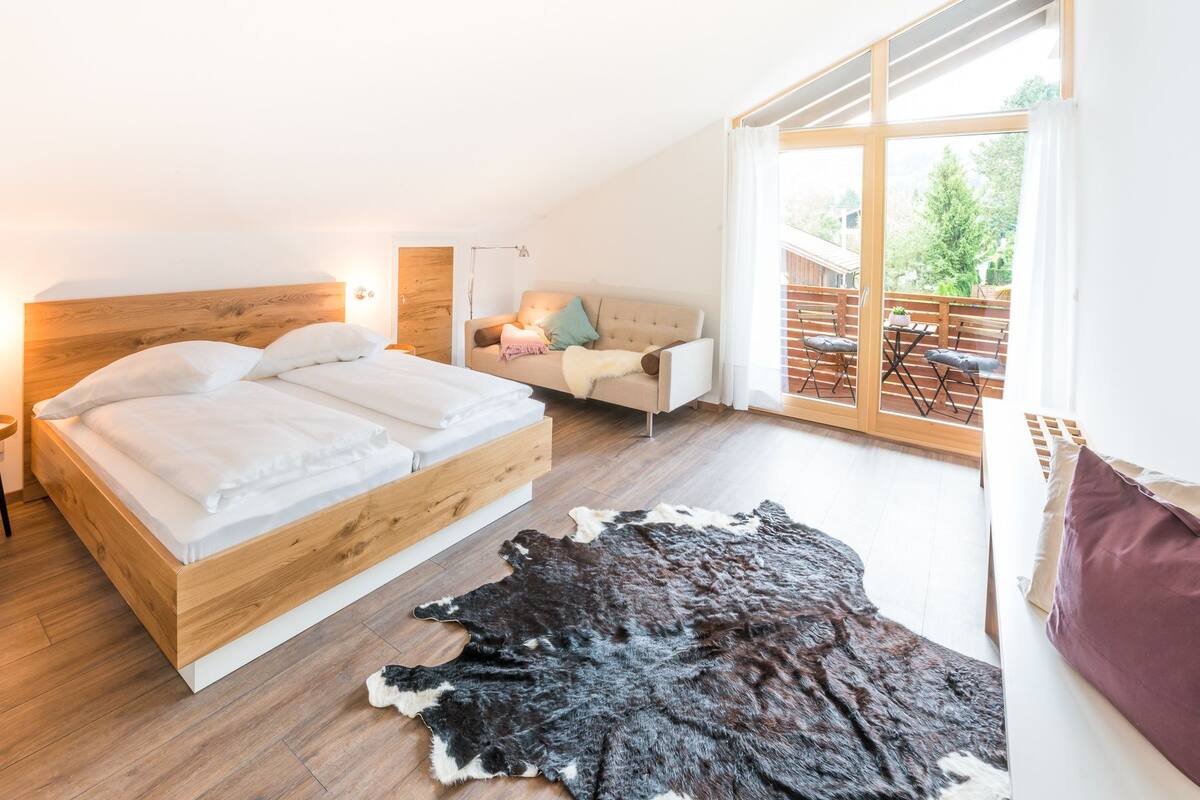 Un dormitorio con una cama de madera, una mesita auxiliar pequeña, un sofá, un ventanal grande y una alfombra de cuero.