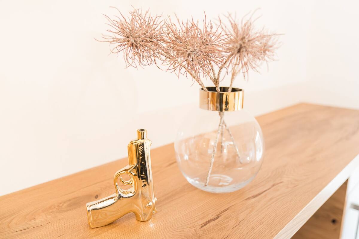 Una mesa de madera con un adorno dorado y un florero de vidrio transparente con plantas secas.