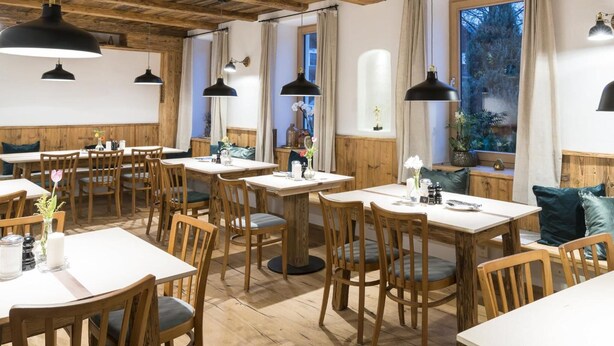 A restaurant with wooden tables and chairs, pendant lighting, and large windows with curtains. - Stefanies Cafe Pension Culture (Bad Feilnbach)