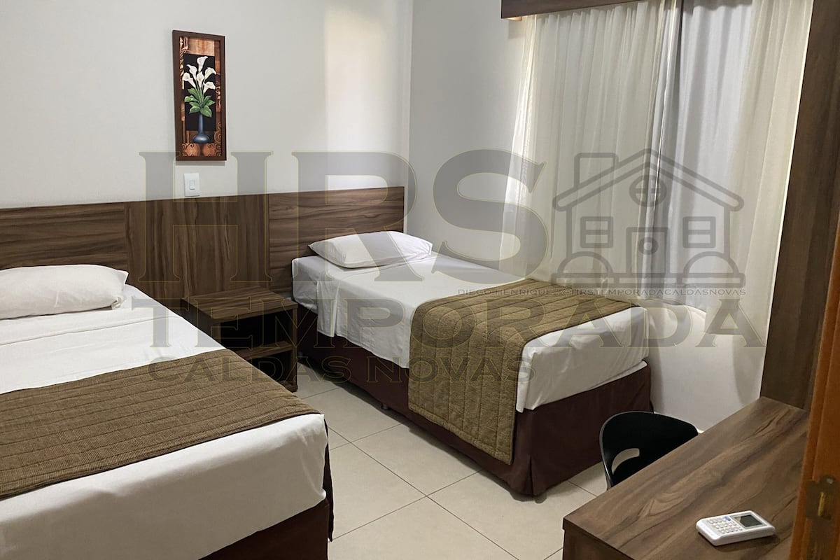Ein Hotelzimmer mit zwei Einzelbetten, einem Holzkopfteil, einem kleinen Nachttisch, einem Schreibtisch mit Stuhl und einem Vorhangfenster.