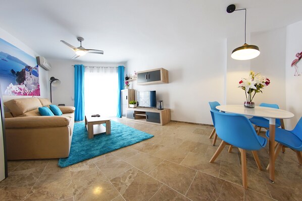 A modern living room with a sofa, coffee table, and blue chairs. There is a dining table with a vase of flowers. The room has a ceiling fan, a TV, and a cabinet. - Zenplaya bajo 8 Playa Bajondillo (Torremolinos)