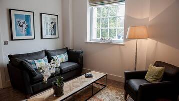 Un salon avec un canapé en cuir noir, une table basse, une table d’appoint, une lampe et deux tableaux encadrés accrochés au mur.