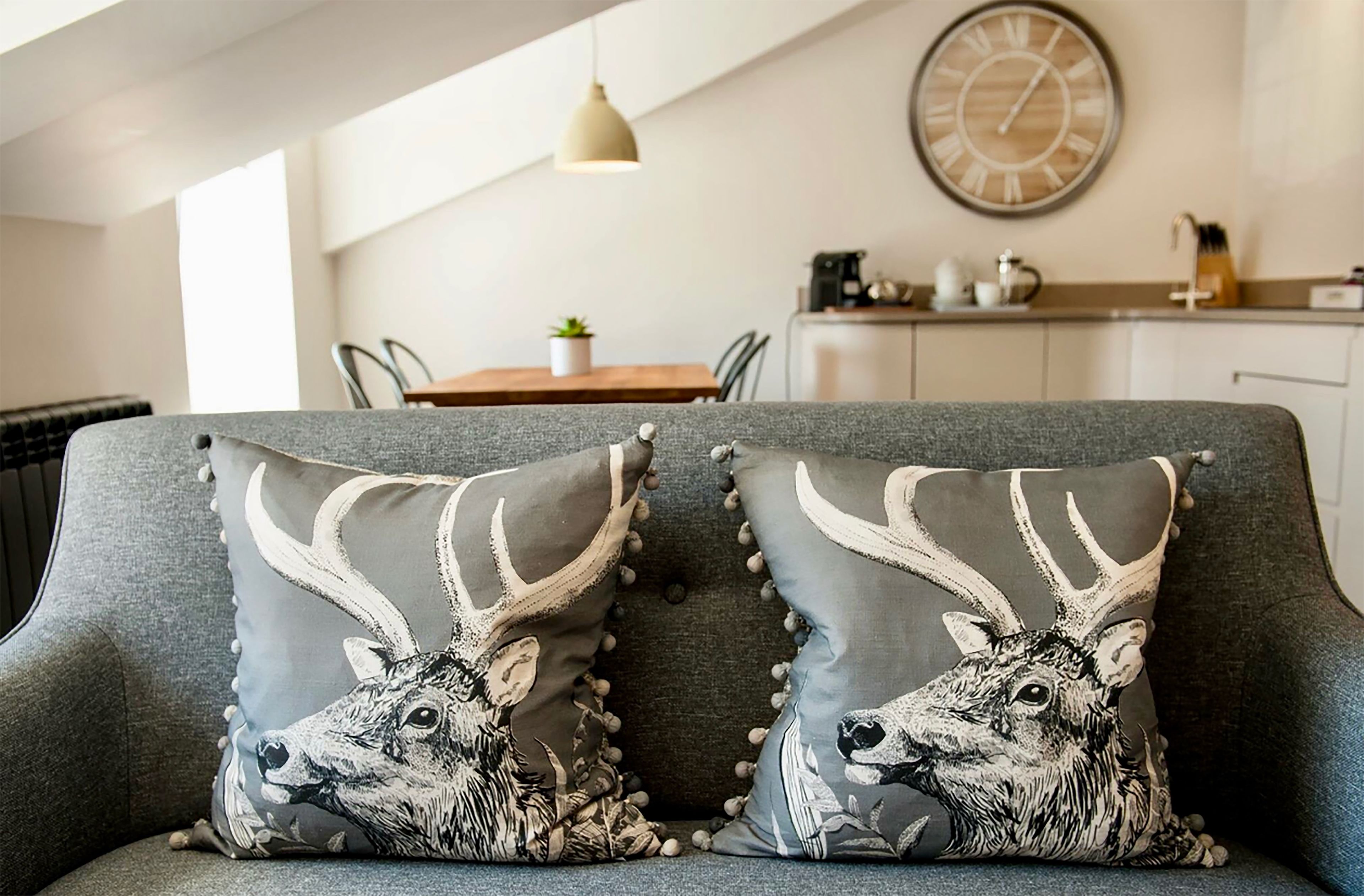 A modern living room with a grey sofa, two decorative pillows with deer designs, a round wall clock, and a kitchen area in the background.
