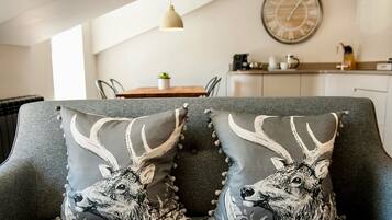 A modern living room with a grey sofa, two decorative pillows with deer designs, a round wall clock, and a kitchen area in the background.