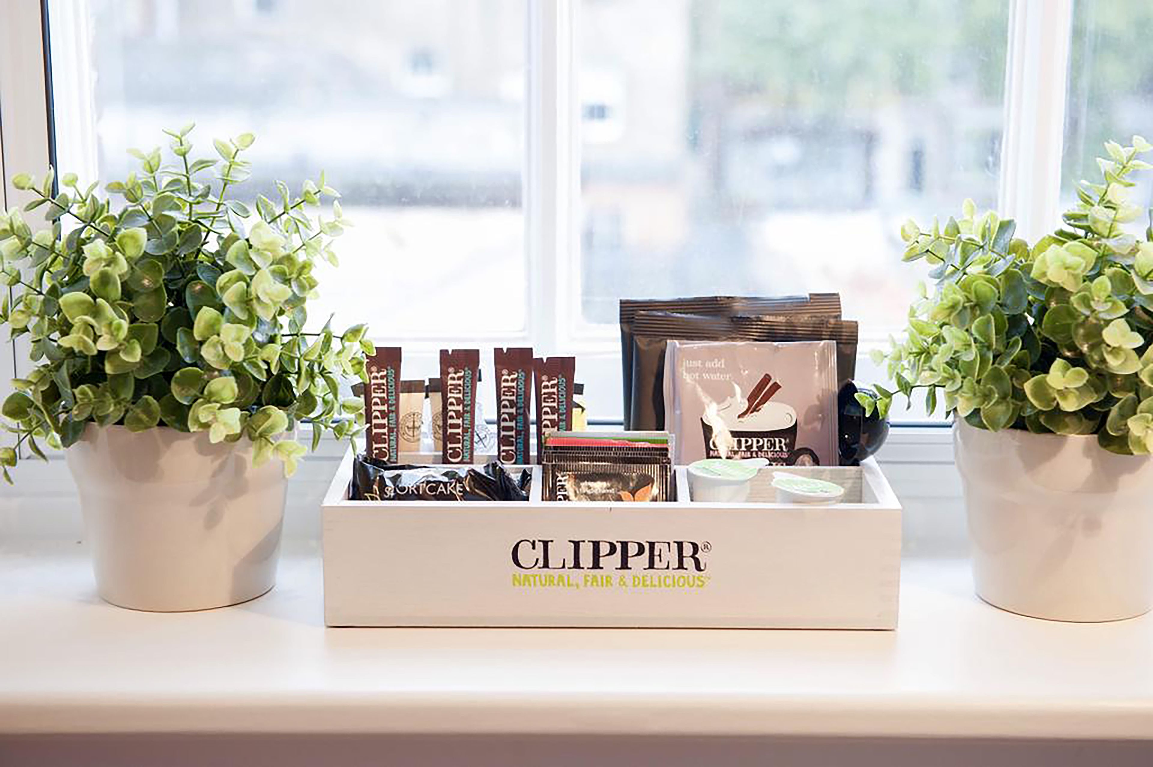 Egy tálca Clipper márkájú termékekkel az ablakpárkányon, mellette két virággal ültetett növény.