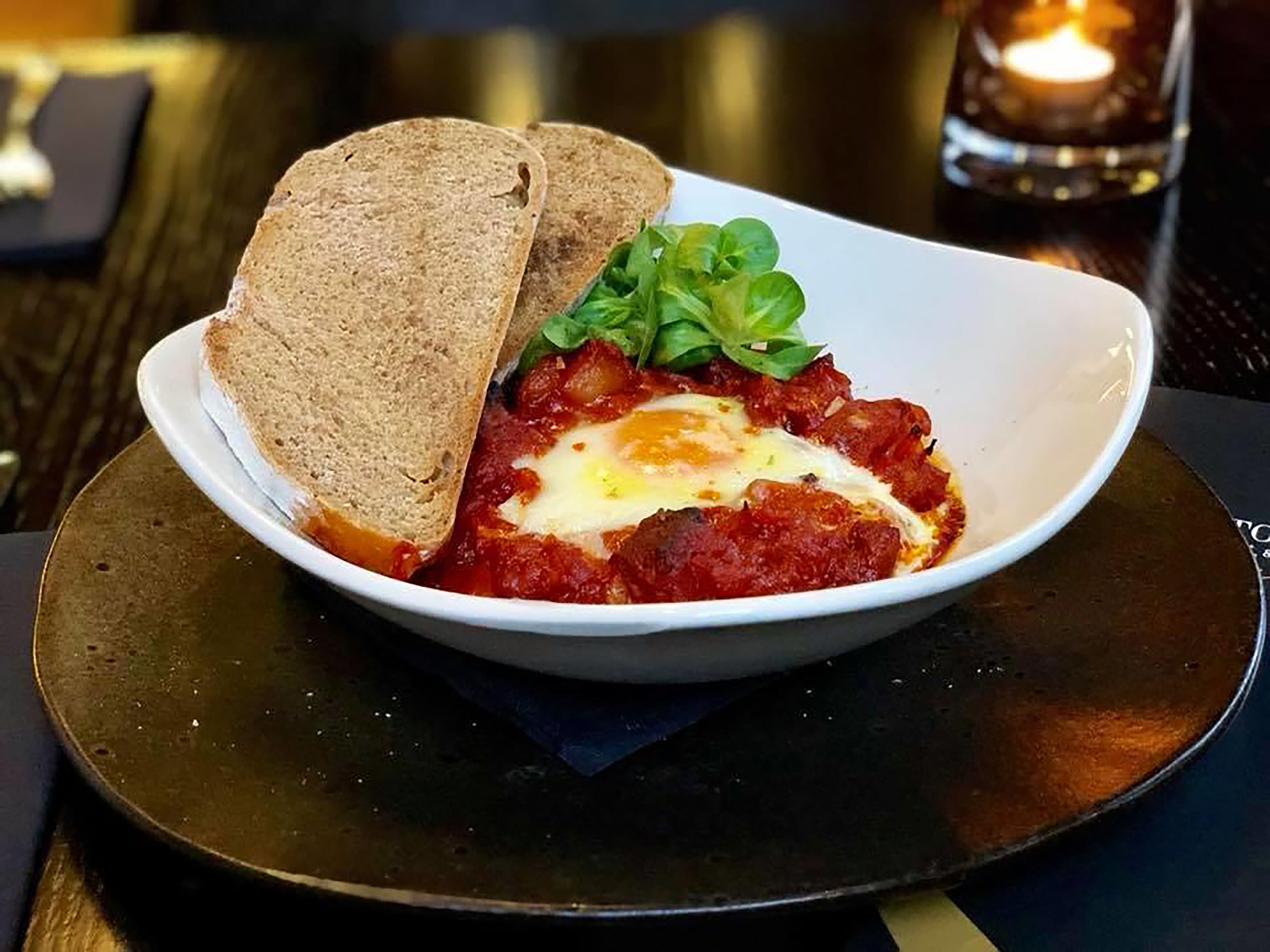 A bowl of shakshuka with toast and greens on a plate.