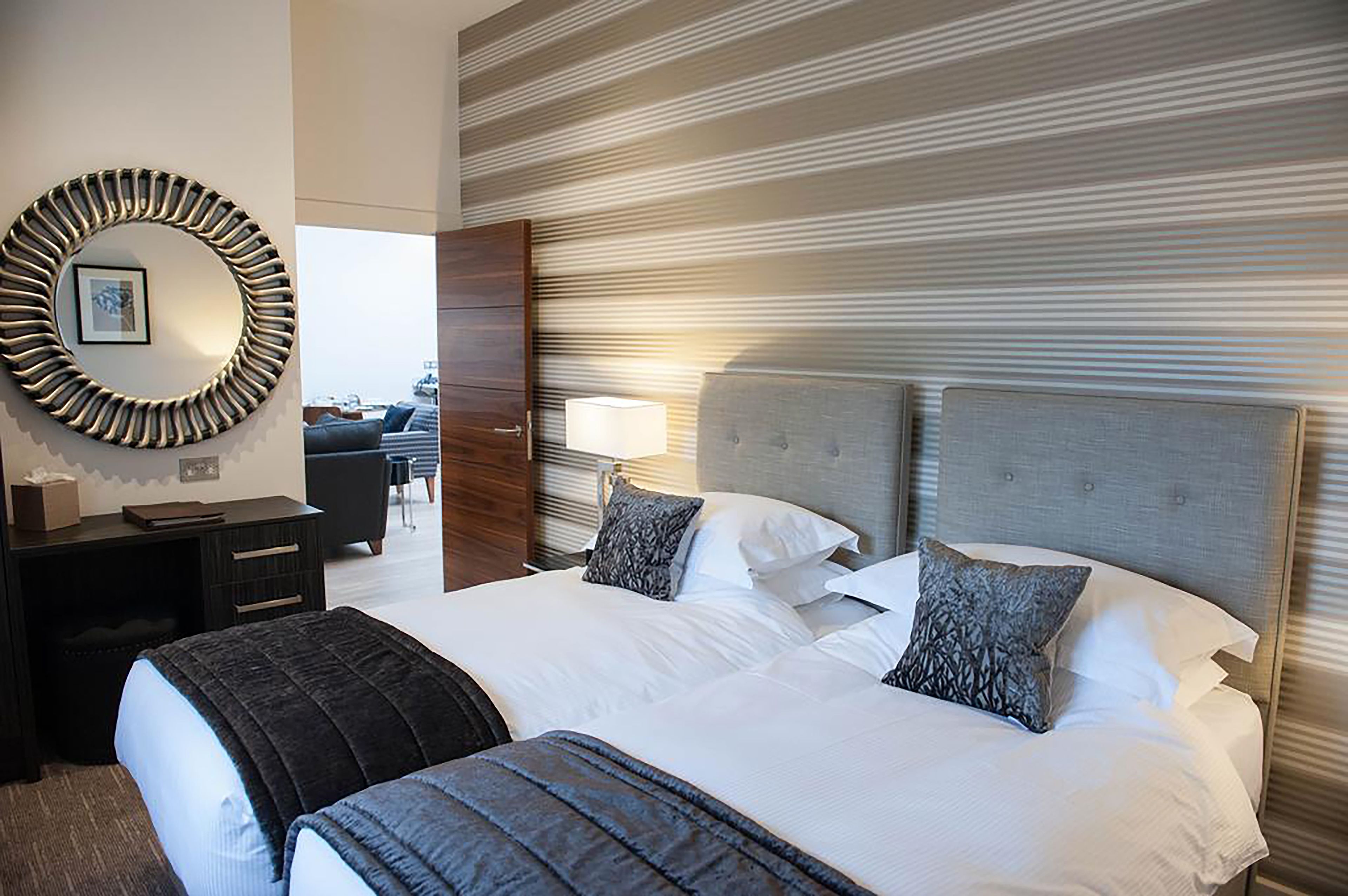A hotel room with a large bed, a round mirror, a desk with a chair, and a view of the city through the window.