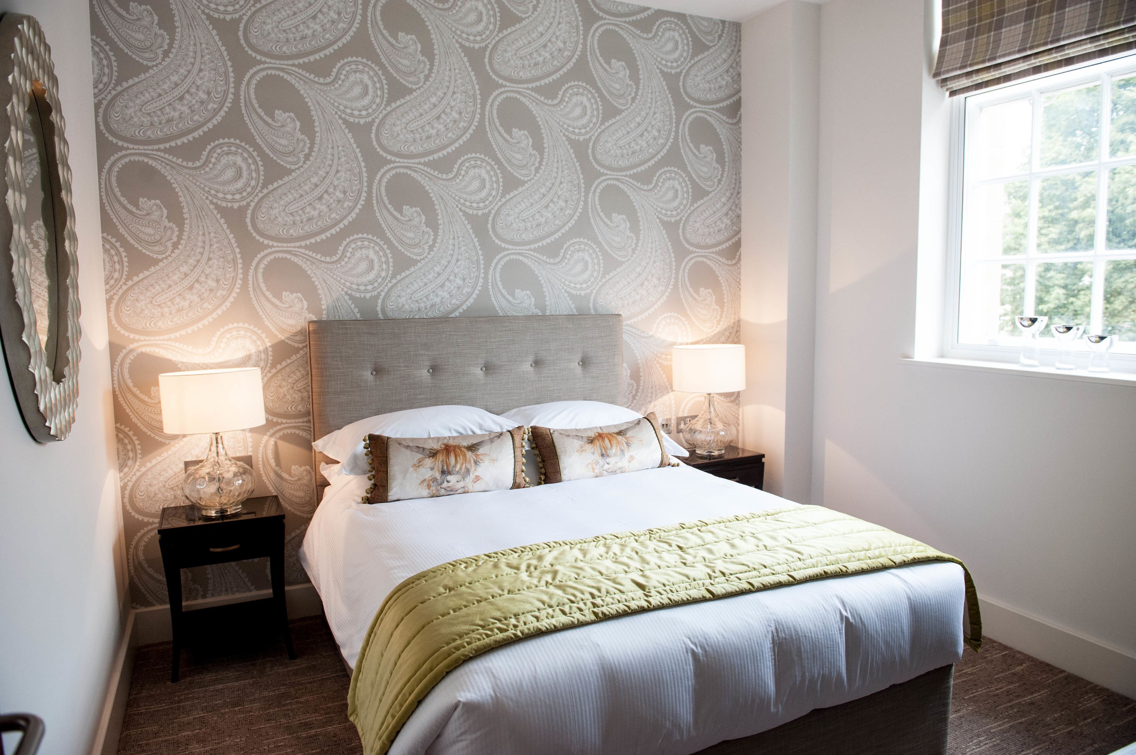 A neatly made bed with a green blanket, two bedside lamps, and a patterned wallpaper.