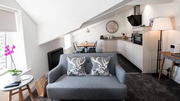 En moderne stue med en grå sofa, et spisebord med stole, et tekøkken og et fjernsyn ophængt på væggen.