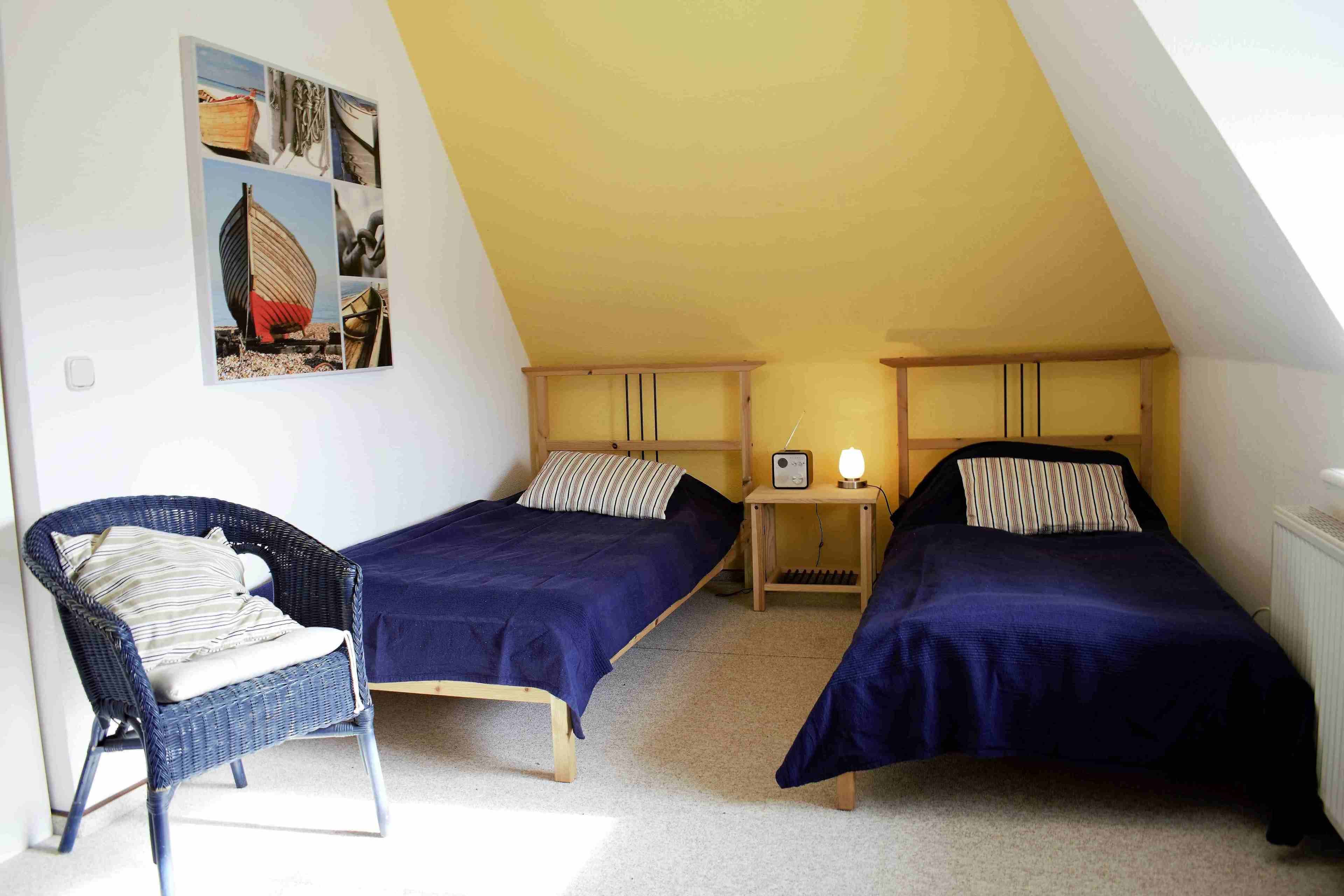 Ein kleines, gemütliches Schlafzimmer im Dachgeschoss mit zwei Einzelbetten, einem Korbstuhl und einem Wandbild.