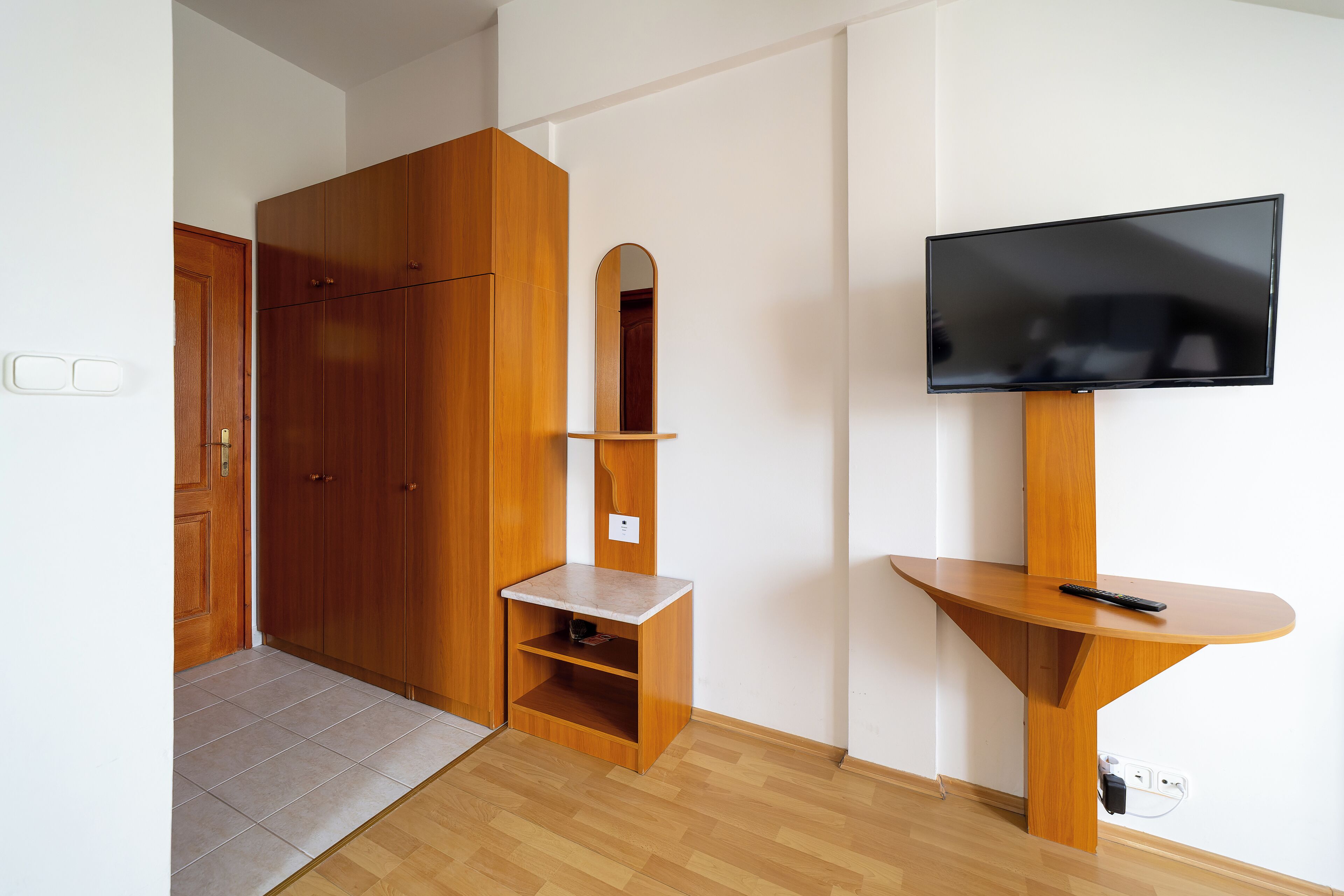 Habitació amb armari de fusta, una petita estant, televisió muntada sobre un suport de fusta i terra amb rajola.