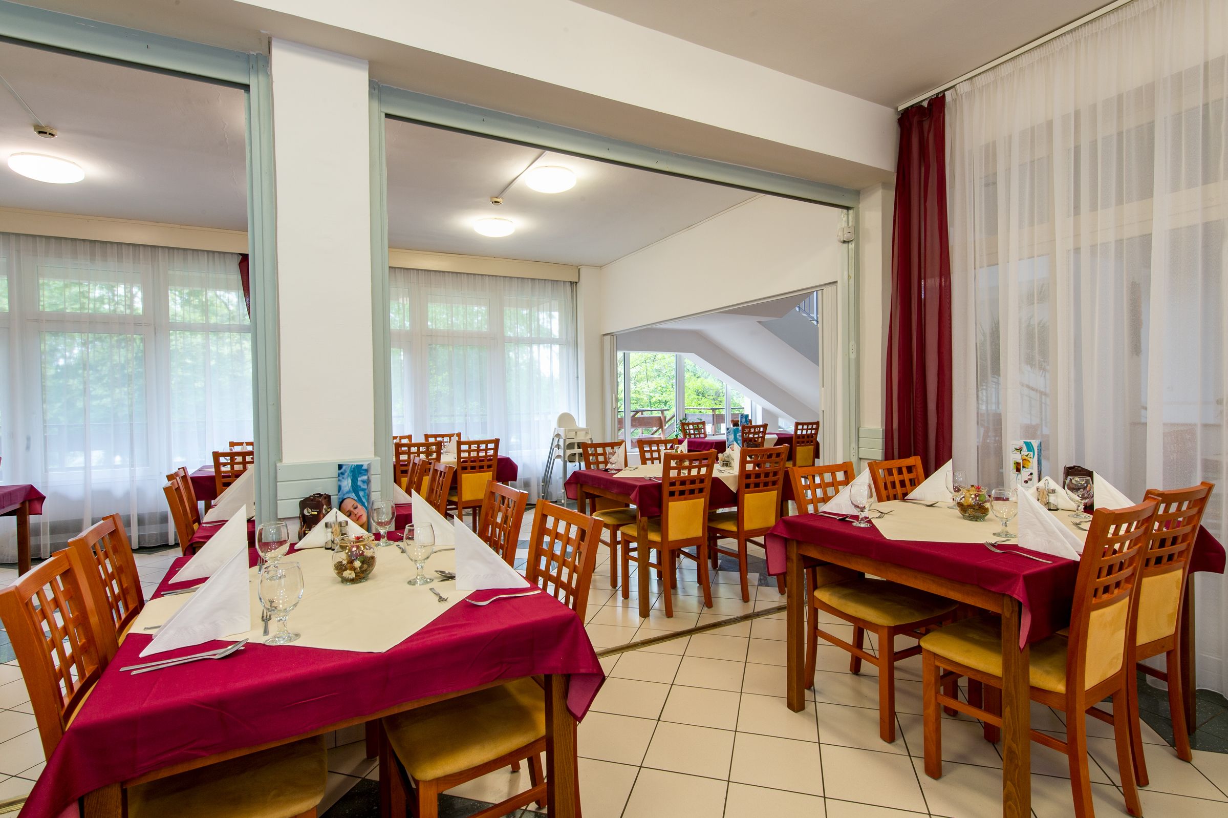 A dining area with multiple tables and chairs, large windows with curtains, and a view of greenery outside.