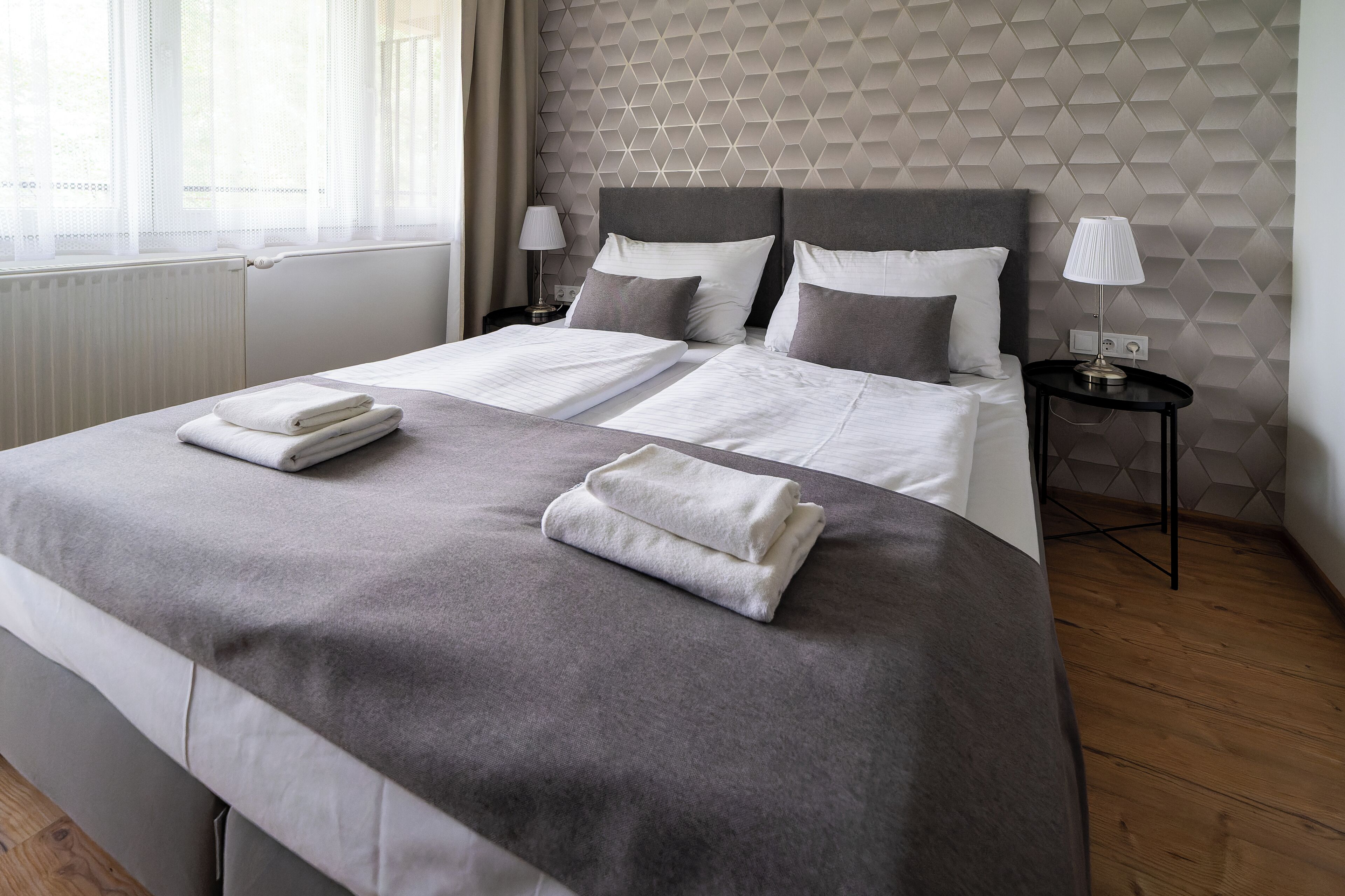 A neatly made bed with two pillows and folded towels, a bedside table with a lamp, and a geometric-patterned wall.