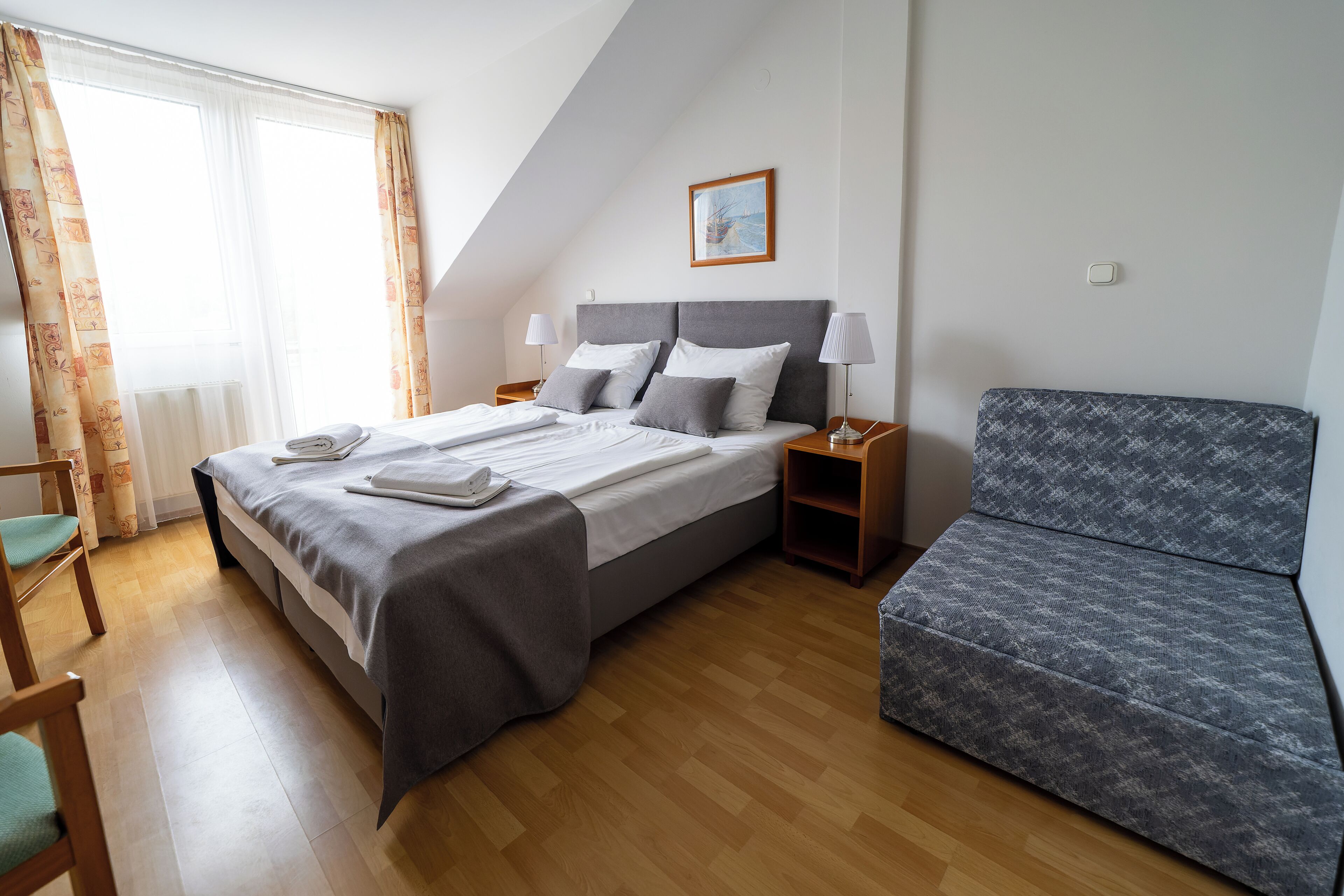 A hotel room with a bed, two bedside tables, a chair, a sofa, and a window with curtains.