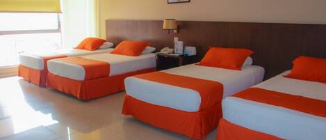 A hotel room with four beds, each with orange bedding and white pillows. There is a nightstand with a lamp and a phone, and a window with a yellow blind.