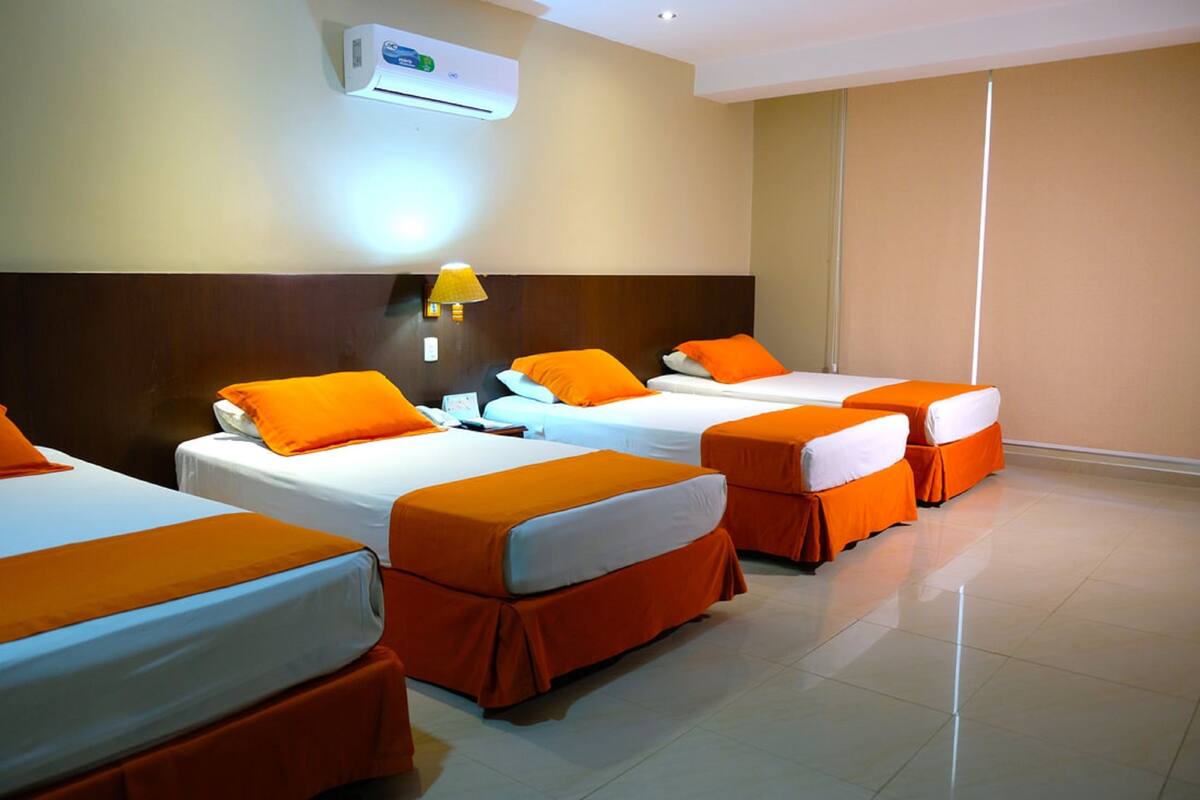 Habitación de hotel con cuatro camas dispuestas en fila, cada una con ropa de cama naranja y almohadas blancas.