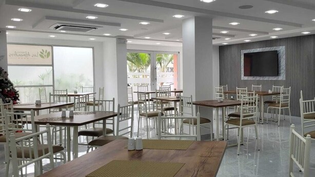 A restaurant with white chairs and tables, a large window, and a flat-screen TV mounted on the wall. - Hotel Los Almendros (Manta)