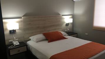 A hotel room with a bed, a nightstand with a phone, a wall-mounted lamp, and an air conditioning unit.
