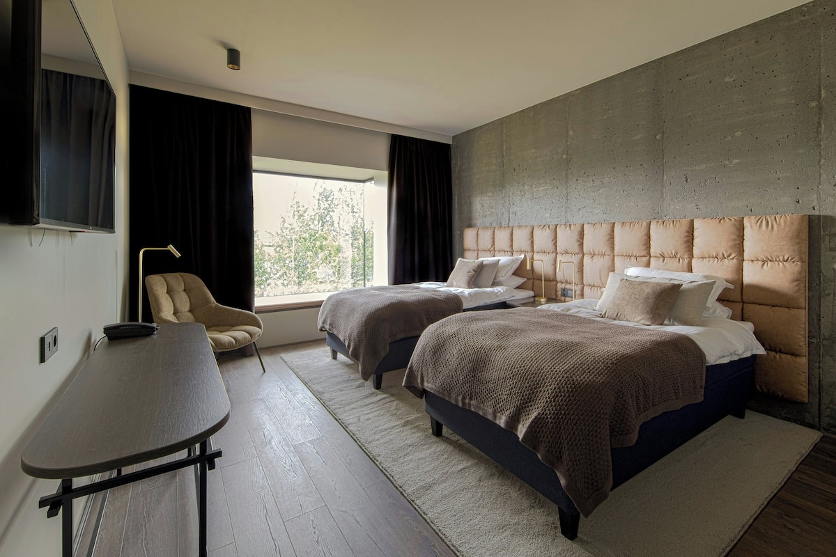 A hotel room with two beds, a desk, a chair, a large window, and a concrete accent wall.