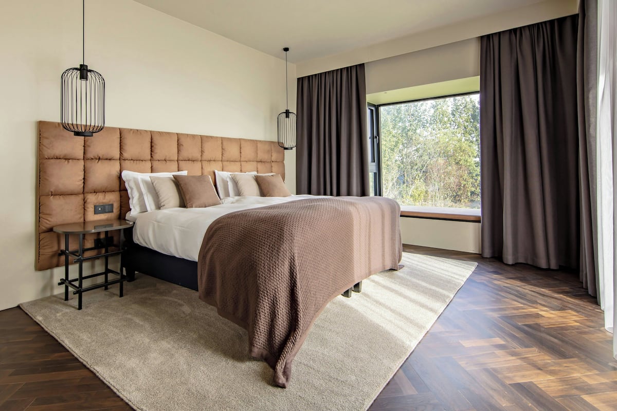 A bedroom with a large bed, a bedside table, a hanging light fixture, and a view of the outdoors through a window.