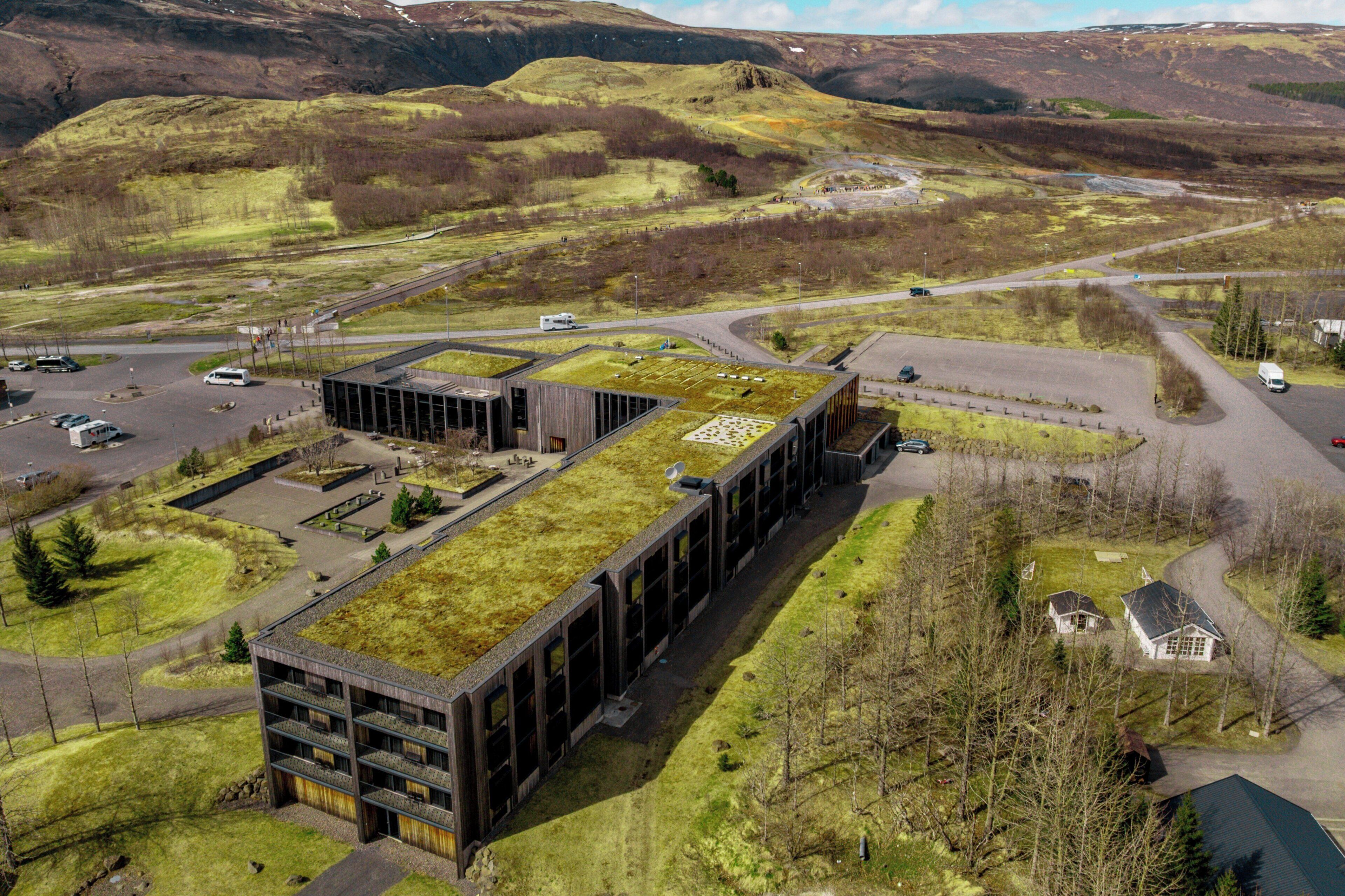 Vista aérea de un edificio moderno con techo verde, rodeado de un estacionamiento y un paisaje montañoso.