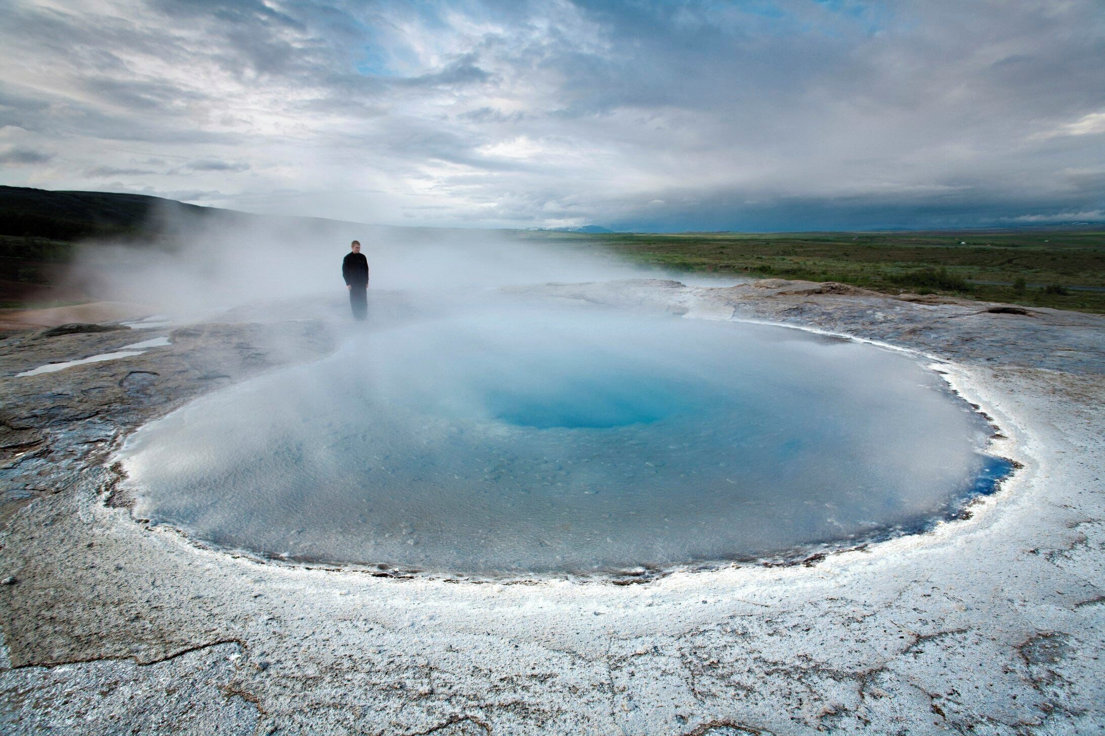 Una persona de pie en una piscina geotérmica con vapor que se eleva, rodeada por un paisaje árido.