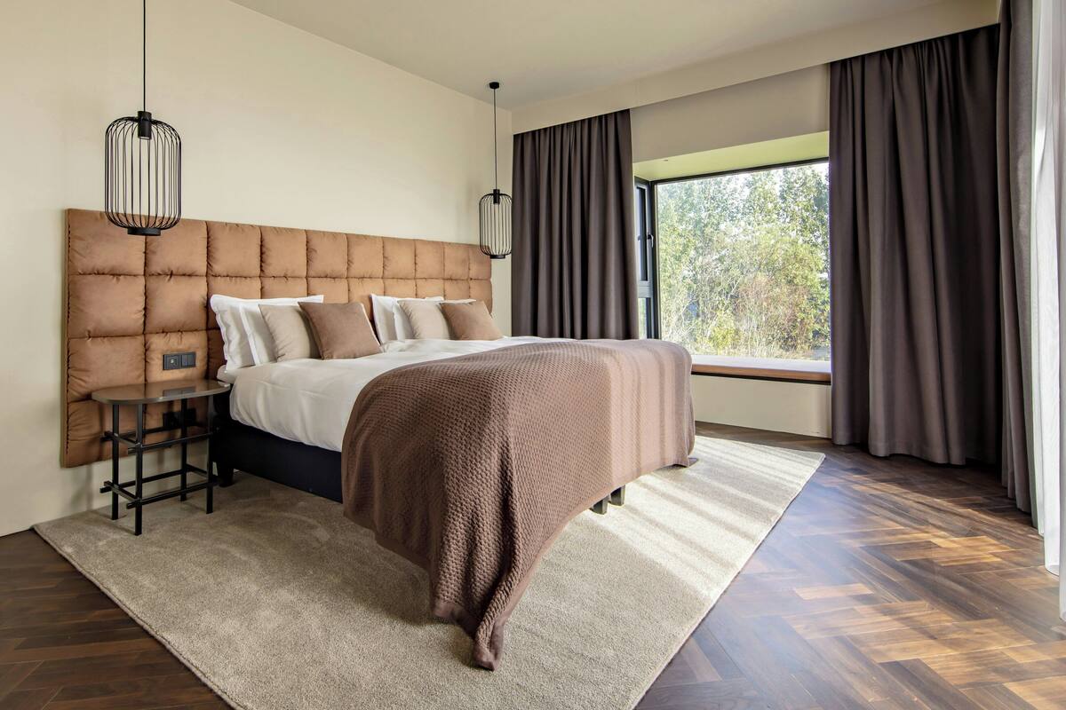 A bedroom with a large bed, a bedside table, a hanging light fixture, and a view of the outdoors through a window.
