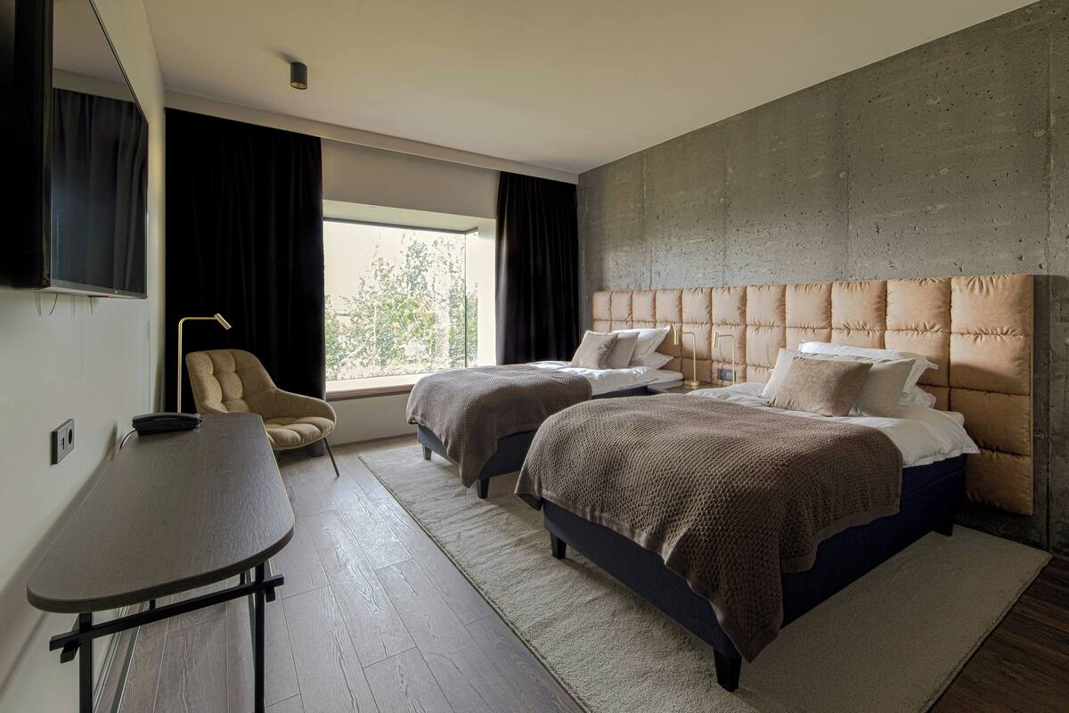 A hotel room with two beds, a desk, a chair, a large window, and a concrete accent wall.