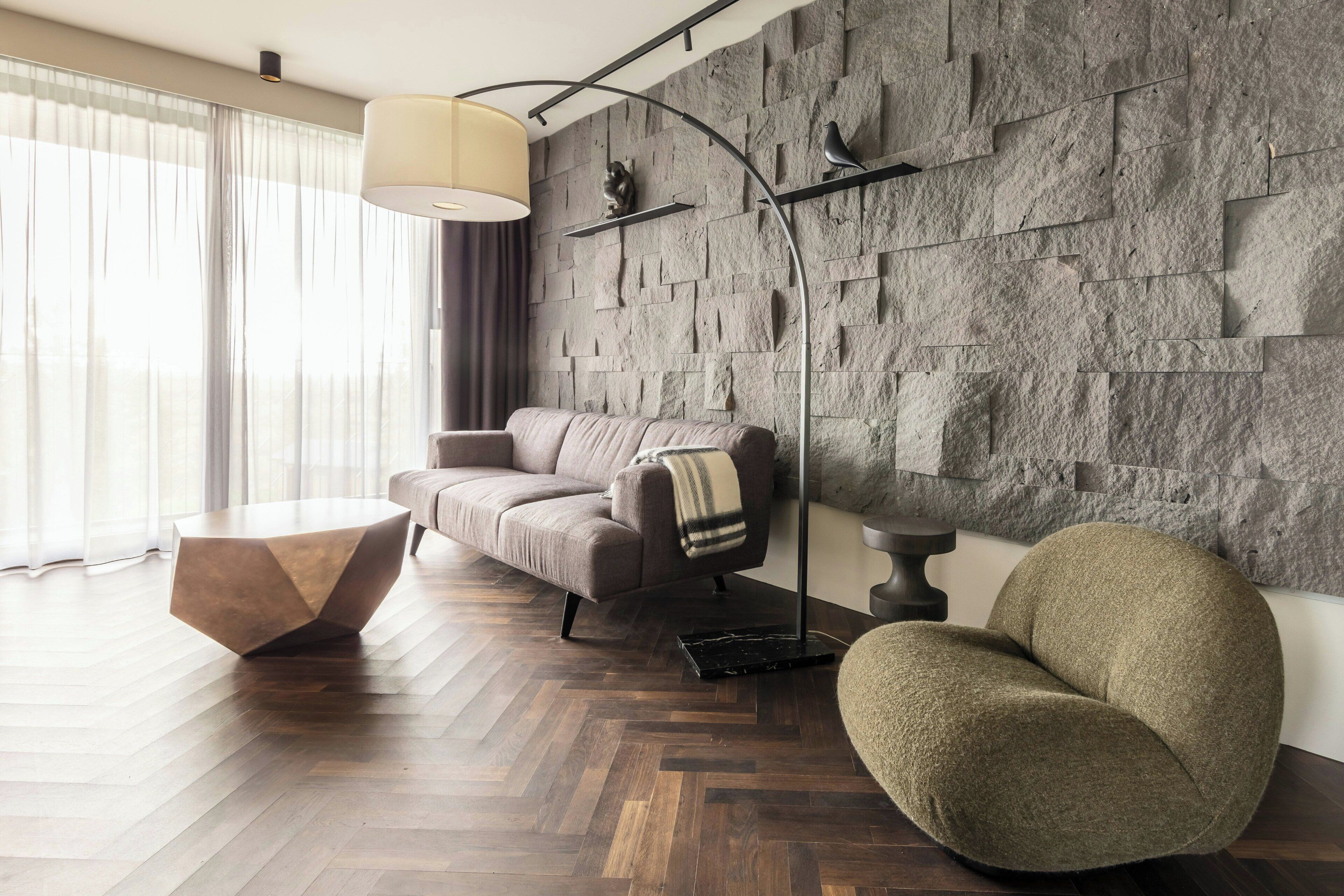 Un salon moderne doté d’un parquet en bois à motifs chevrons, d’une table basse ronde, d’un canapé, d’un fauteuil rond et d’un mur en pierre orné d’une sculpture d’oiseau.