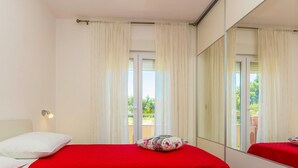 Un dormitorio con una colcha roja, un almohadón blanco, un cojín decorativo, una lámpara de noche y un espejo grande que refleja la habitación y un balcón con vegetación.