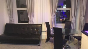 A living room with a black leather sofa, a glass dining table with four chairs, and a view of the city at night through large windows. - Paisley central luxury Rooms & Apartment (Paisley)
