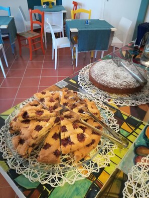 Un coin repas avec une table mise pour le repas, où l’on aperçoit une tarte et un gâteau posés sur un chemin de table en dentelle.