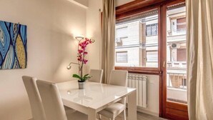 A dining area with a white table and chairs, a potted orchid, and a view of a building through a window with a wooden frame. - Retro2 Vaticano-max 4 pax (Rome)