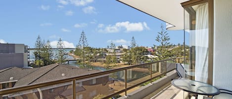 Un balcon doté d’une balustrade en verre, d’une table et de chaises, donnant sur un quartier résidentiel avec des maisons et des arbres.