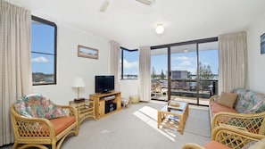 A spacious living room with a large television, comfortable seating, and a balcony with a view. - Sundial 602, Hollingworth Street (Port Macquarie)
