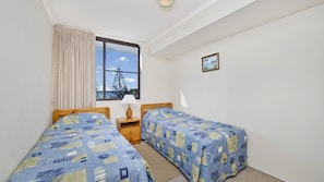 A bedroom with two beds, a window with a view of trees, a bedside table with a lamp, and a framed picture on the wall. - Sundial 602, Hollingworth Street (Port Macquarie)