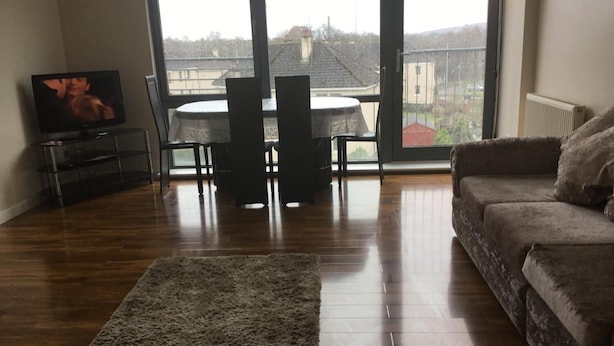Ein Wohnzimmer mit Holzboden, einer Couch, einem Fernseher, einem Esstisch mit Stühlen und Blick durch große Fenster.