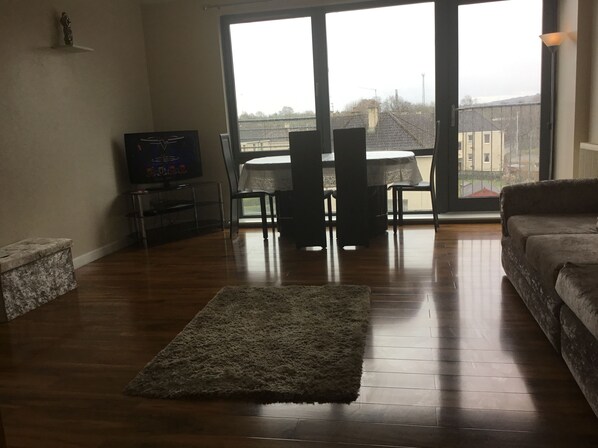 A living room with a wooden floor, a grey rug, a sofa, a TV on a stand, and a dining table with chairs.