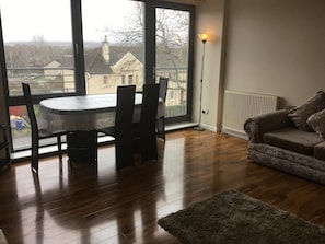 A room with a dining table and chairs, a sofa, a radiator, and a large window with a view of houses and trees.
