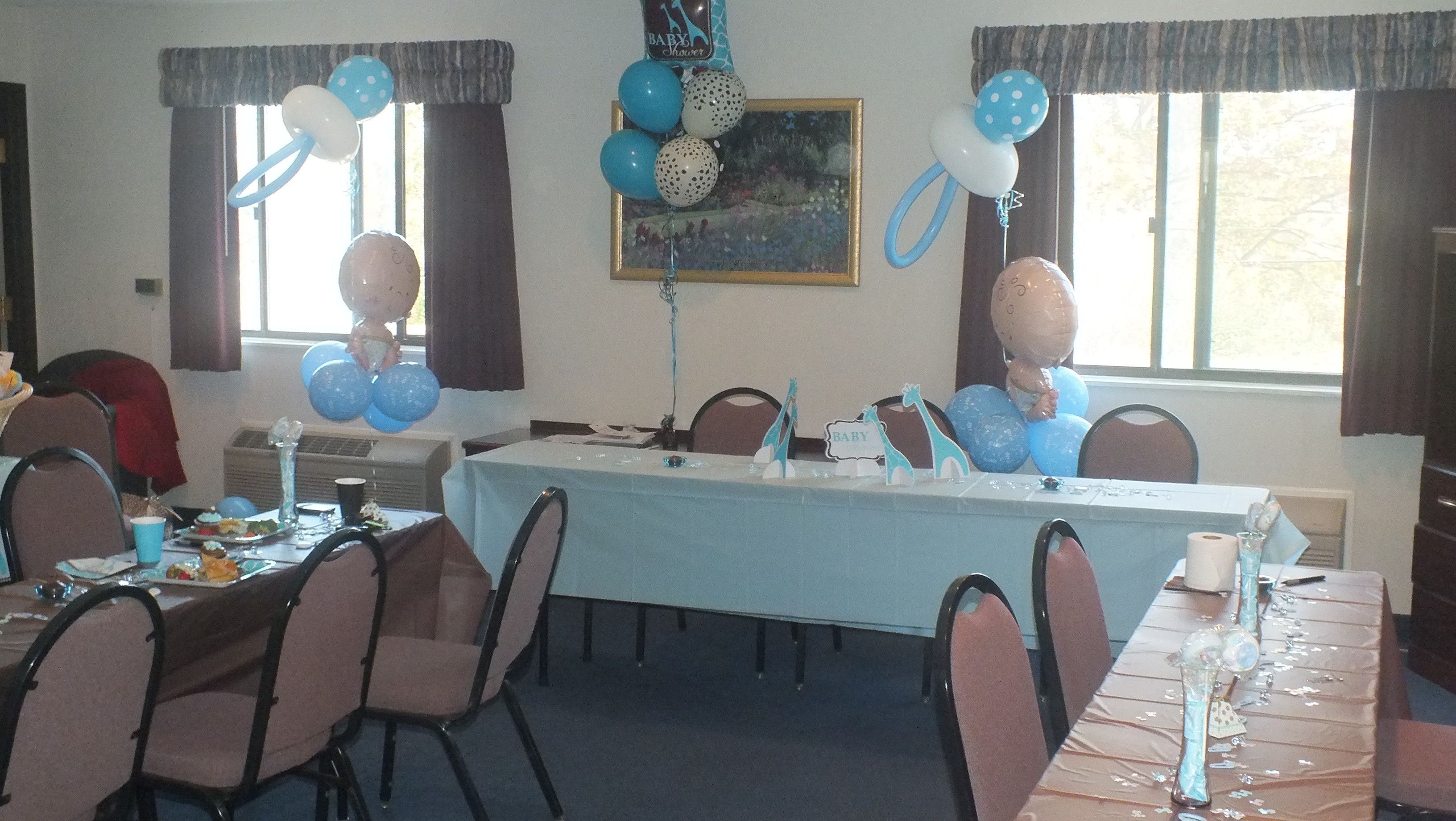 A room with a long table set for a baby shower, decorated with balloons and a painting on the wall.