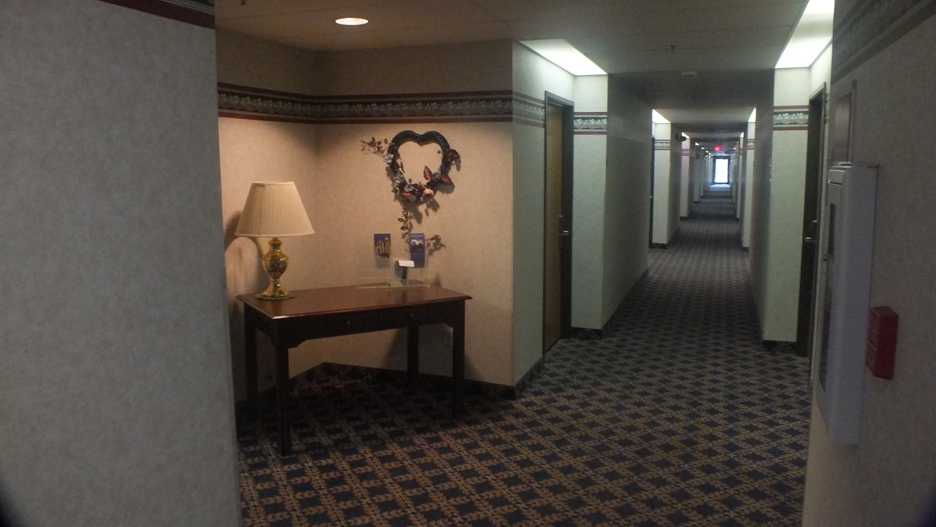 A hotel hallway with patterned carpet, a table with a lamp, and a decorative wall piece.