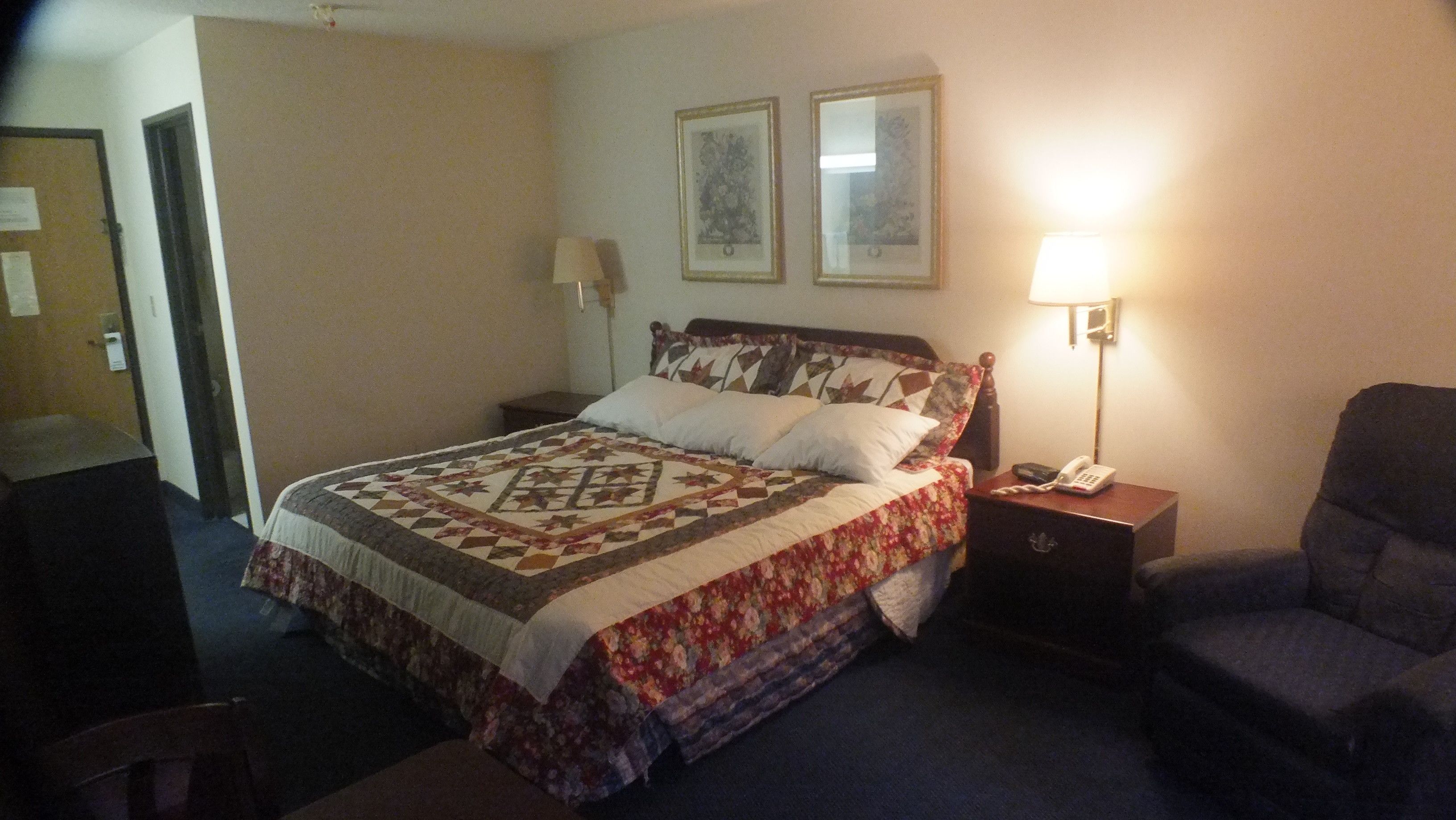 A hotel room with a bed, a nightstand with a phone, a chair, and a lamp.