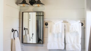A bathroom with a mirror, two wall-mounted light fixtures, and two towels hanging on a towel rack. - Cliffside Tiny Home Container  #5 (Orderville)