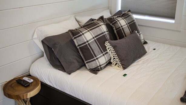 A neatly made bed with a white comforter and multiple pillows, including a plaid one. A remote control is on a small wooden side table beside the bed. - Cliffside Tiny Home Container  #5 (Orderville)