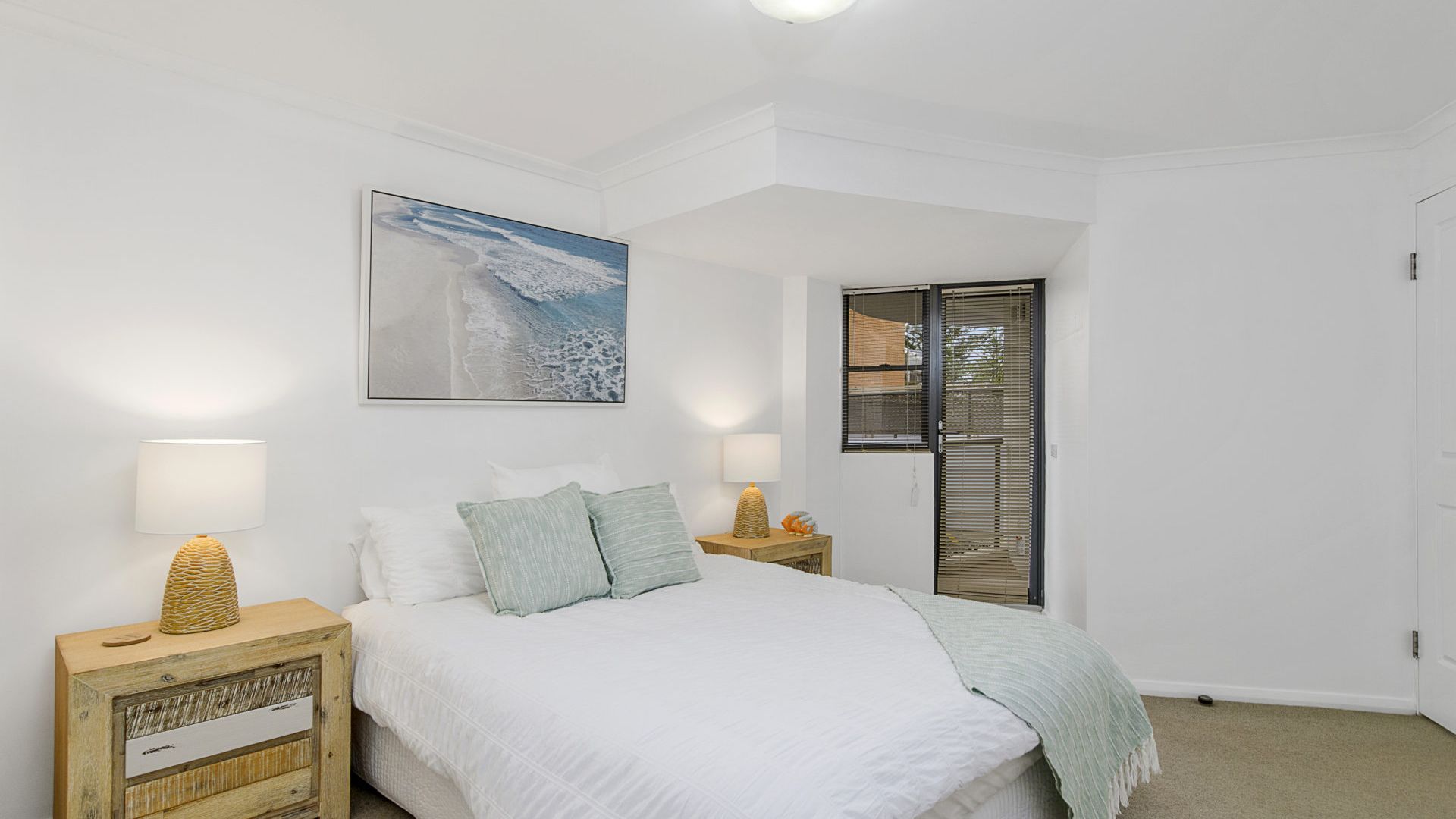 Une chambre à coucher avec un lit, deux tables de chevet avec des lampes, un tableau accroché au mur et une porte avec des stores.