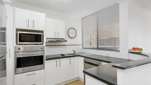Een moderne keuken met witte kasten, roestvrijstalen apparatuur, een zwart aanrecht en een raam met jaloezieën.