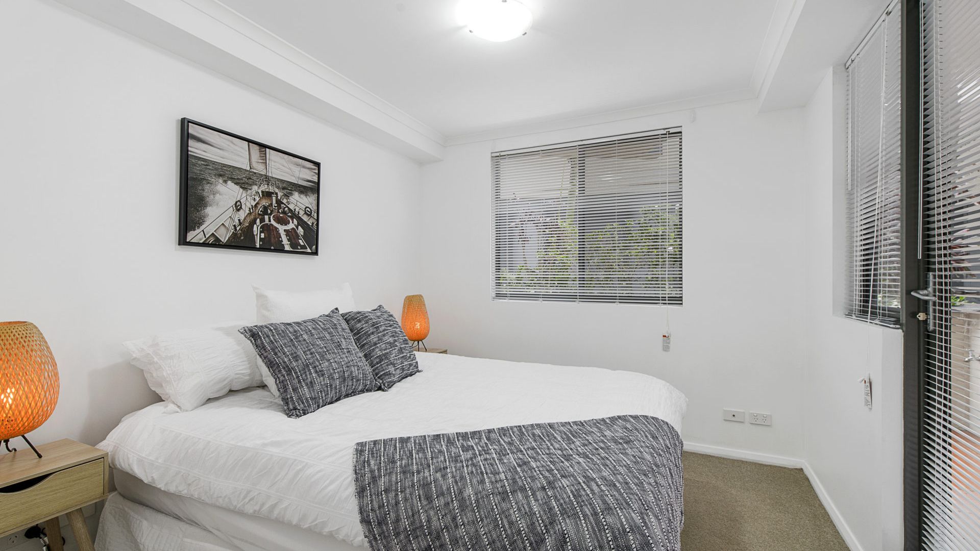 Ein Schlafzimmer mit einem Bett, einem Nachttisch, einer Lampe, einem Fenster mit Jalousien und einem an der Wand hängenden Bild.