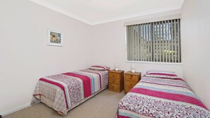 A bedroom with two single beds, a small wooden nightstand, a nightstand lamp, a framed picture on the wall, and a window with vertical blinds.
